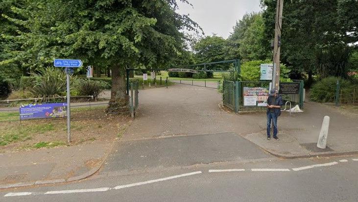 Entrance to a park from a road with green gates either side and a metal green barrier across the top to restrict vehicle height. A man stands to the right of the entrance on the pavement, looking at his phone. There are signs either side of the entrance