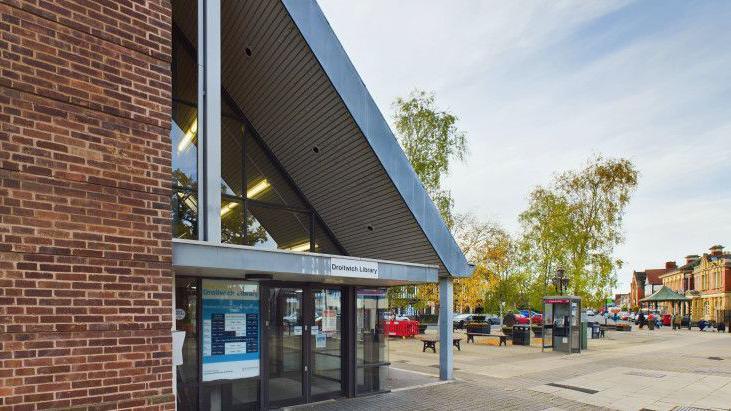 A red brick building with a long sloped metal roof. A sign says Droitwich Library