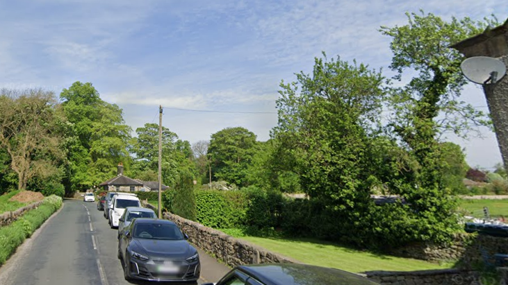 A queue of parked cars along a narrow, rural road. A pavement and dry stone wall are to the side of the cars. Some trees and shrubs are in the background.