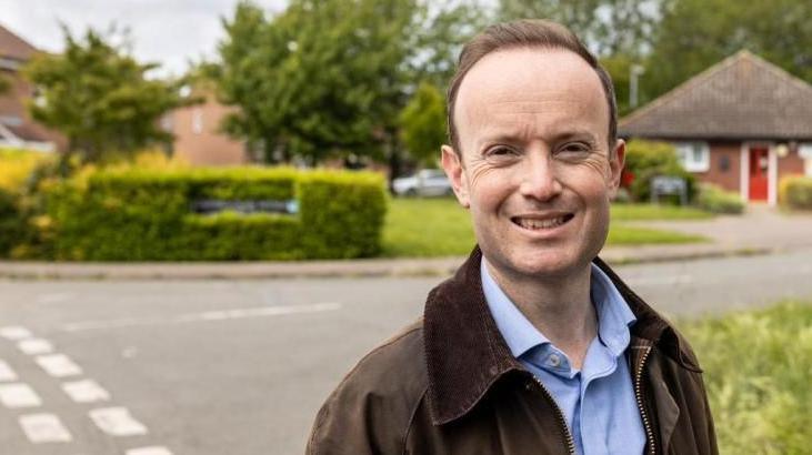 Richard Rout is standing on a street corner next to a grass verge. He is wearing a brown jacket and blue shirt. He is smiling at the camera. In the background you can see a blurred bungalow.