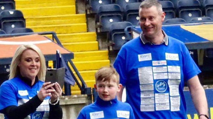 A man, woman and young boy walk around the side of a football pitch while smiling - they are wearing blue tops with lots of names and writing on them, possibly sponsors