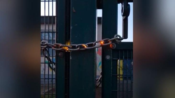 A black metal chain has a orange glow on three of the links. It is wrapped around a green metal fence. Behind the fence is a white brick building with a window. 