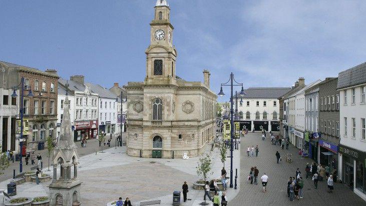 Coleraine town centre. There are shops on either side of the squre and people walking on the street. The sky is blue. 