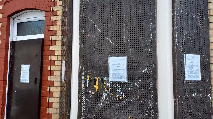 The front window of a brick terrace house has been boarded off with sheets of metal. A4-sized notices have been stuck to the window and front door.