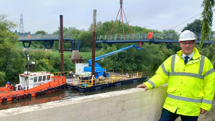 Jon Fraser in a hi-vis jacket and hard white hat standing in front of the construction site of Kepax bridge