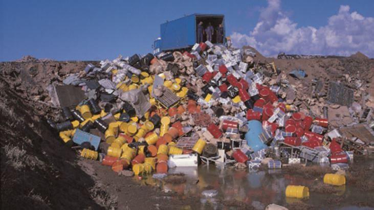 An old photo of a lorry at the top of a trench. Red and yellow metal drums, among other waste, are tipped from the back of the lorry and into the trench