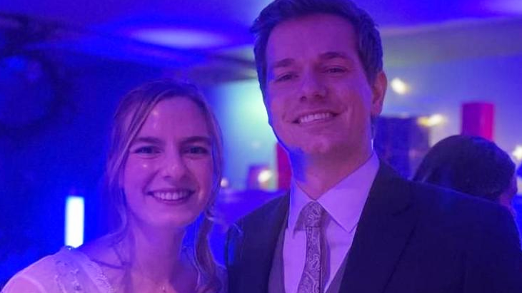 Rebecca and George at the party in wedding dress and suit. She is wearing a white dress with lace sleeves and he is wearing a grey waistcoat and tie and navy suit. Rebecca has long curly blonde hair.