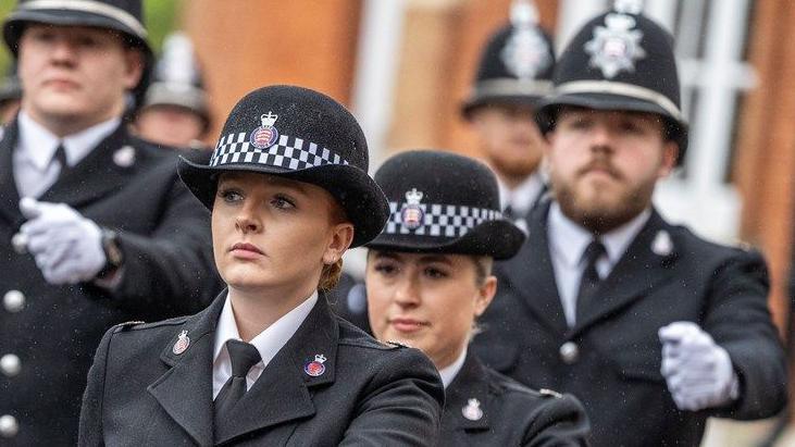 A female police officer marches in front of three others. They are all wearing smart black outfits and hats, with white gloves on.