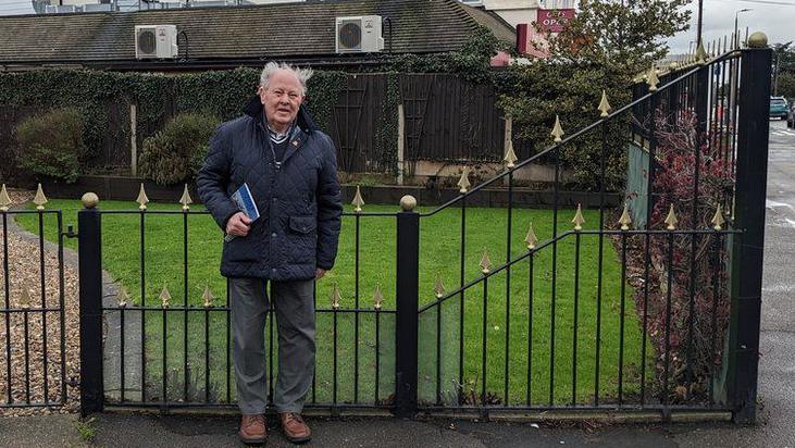 Ray Howard, dressed in a coat and trousers, standing in front of a house with a pebbled driveway, and a car in the background.