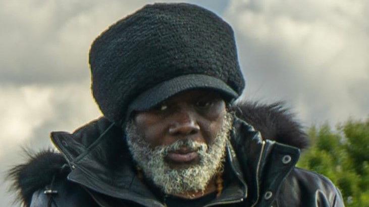 Franklin Francis in a black coat with a hood and a black hat. He has a grey beard. 