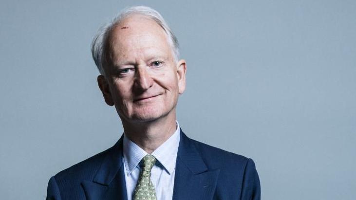 Sir Henry Bellingham has white hair and a mark on his upper forehead. He is smiling. He is wearing a blue jacket, light blue shirt and a green patterned tie. There is a handkerchief in his breast pocket. The background is plain light blue.