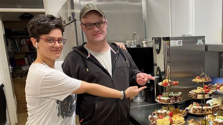 A woman in a white T-shirt and a man in a dark hooded top posing side-by-side and gesturing towards a selection of cakes while standing in a kitchen