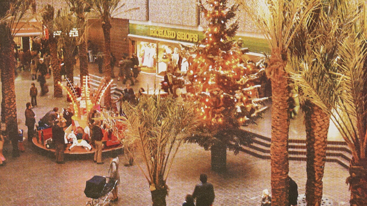 A 1970s picture of a shopping centre full of people with trees scattered around, including a lit-up Christmas tree, with children on a carousel on the left of the frame, next to a shop front in the background that says Richard Shops in yellow writing.