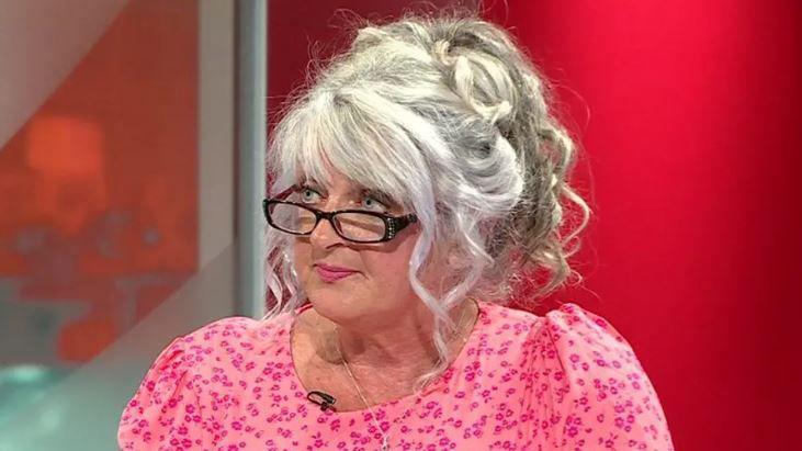 Angie Richardson sits in a TV studio. She has large blonde hair hanging down in curls and is wearing black glasses and a pink floral blouse.