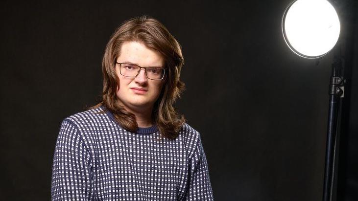 Publicity shot of Dan Bowhay in a dark photography studio. He has long brown hair, is wearing metal-rimmed glasses and a blue and white checked jumper. To the right of the shot is a bright studio light.
