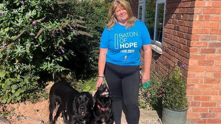 Tracy Ricketts stands outside with her two dogs, wearing a blue Baton of Hope Tour 2025 T-shirt. She is in the garden, next to her house, and there are shrubs behind her and plants in pots.