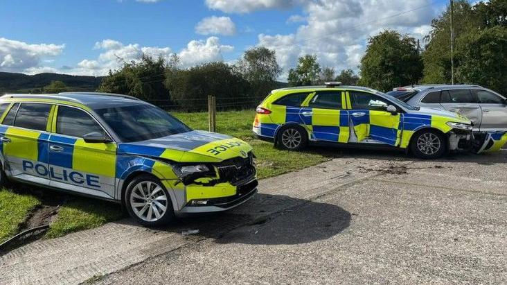 Three wrecked police cars sit partially on some green grass and grey concrete. They all have damage to them. The first, a yellow and blue chequered car, has a wrecked bonnet, with a smashed front grill. The second has a smashed driver side door, it is the same colour as the first. The third is a grey unmarked skoda, with a smashed door panles on the back seat of the driver side. 