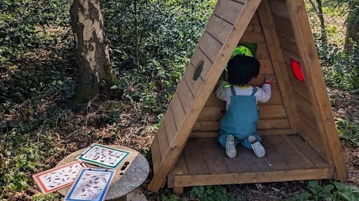A young girl inside a wooden triangular play den. It is outdoors in a forest. A wooden table with cards is next to the den