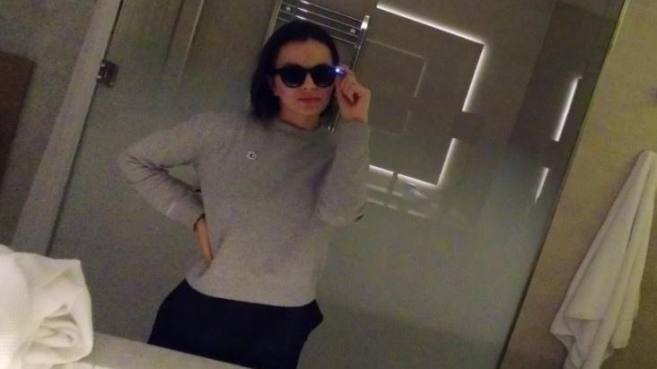 Woman with shoulder length black hair and grey jumper taking a photo of her reflection in the mirror wearing dark glasses. 