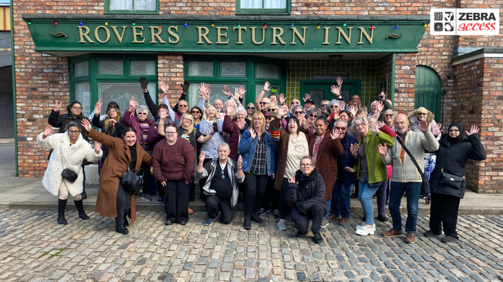 A large group of people stand in a group on the Coronation Street filming set, on the cobbles outside the Rovers Return Inn.