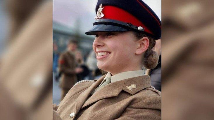 Jaysley Beck wearing her brown formal military uniform and a black and red cap. She has her light brown hair tied back into a low bun and is looking off to the left of the camera and smiling.