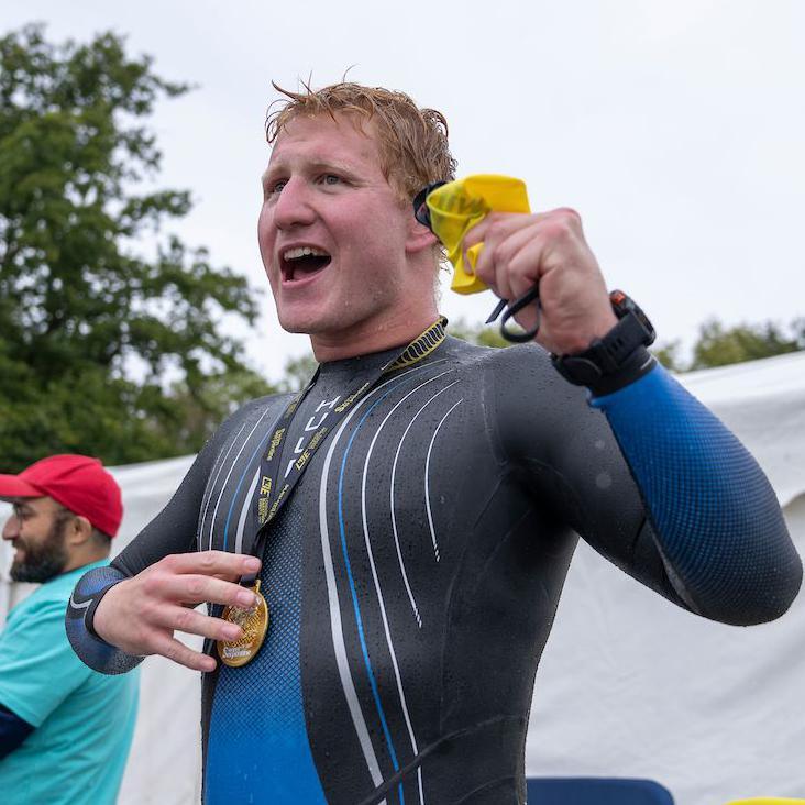 Angus Leckonby in a blue wetsuit with short hair with a medal around his neck. There is a man to the side of him with a blue T-shirt, red cap and beard. There are some trees.