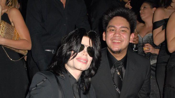 A photograph of pop star Michael Jackson with black hair and aviator sunglasses. Pictured with Prince Azim at his 25th birthday party at Stapleford Park Hall. They are both wearing all black suits
