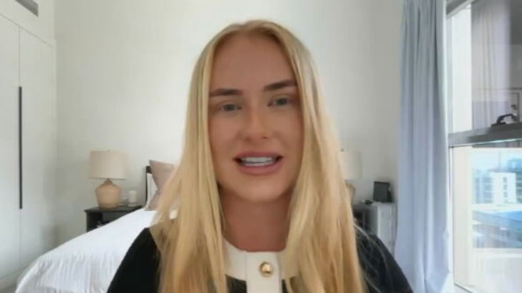A woman with long blonde hair sits indoors in a bright, modern bedroom, speaking toward the camera. She is wearing a black top with a white collar and gold buttons. Behind her is a neatly made bed, bedside table with a lamp, built-in wardrobes, and a large window with curtains showing nearby buildings outside.