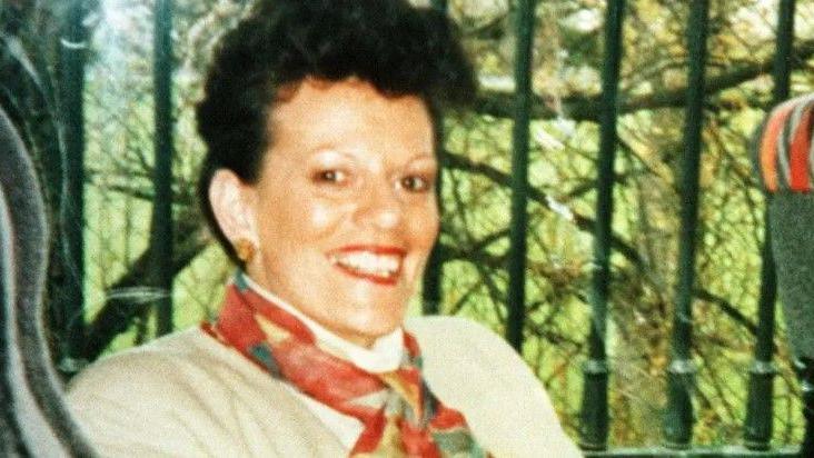 A woman sitting on a coach smiling at the camera. She is wearing a red scarf and tan blazer. She has short black hair. Outside the window of the coach there are green railings and behind that some trees.