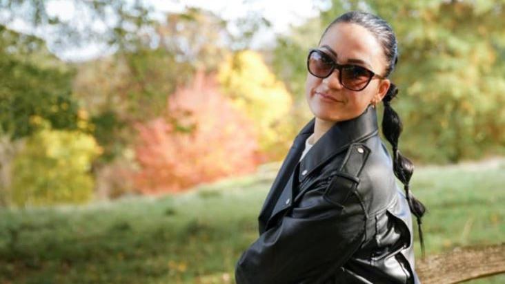 Close-up of Annette wearing dark glasses and a leather jacket sitting in a park and looking to camera