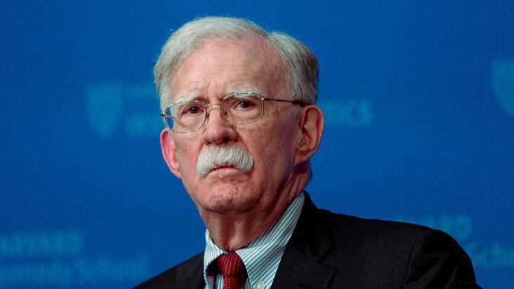 A close-up image of John Bolton, who is looking straight ahead. He is wearing glasses, a black blazer, a stripped blue and white shirt and a red tie.