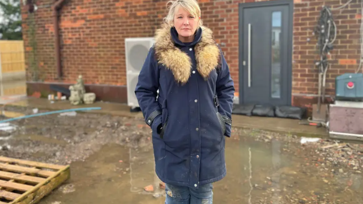 A woman with blonde hair stands in a navy winter coat and wellies surrounded by dirty water.