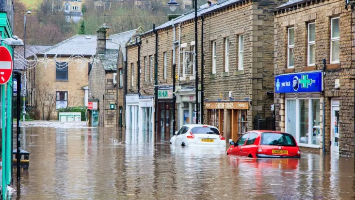 A street in Holmfirth in flood water with two partially submerged cars parked outside a pharmacy