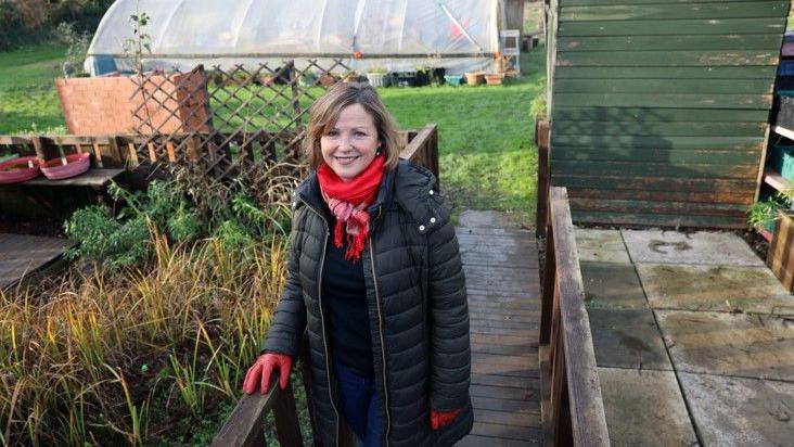 Councillor Karen Clark at an allotment. She has short blonde hair and is wearing a dark green puffer jacket, black top nad jeans, with red gloves and scarf.