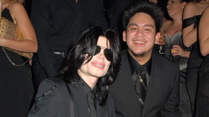 Michael Jackson with long hair and dark aviator sunglasses poses with a grinning man in a black suit, black shirt and black tie with partygoers in the background