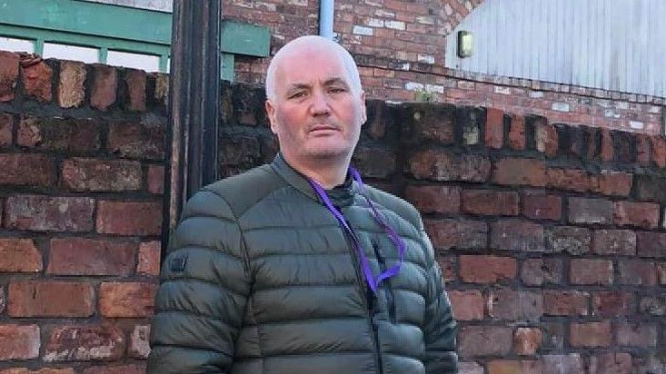 Gary Nicol leaning against a brick wall wearing a green jacket and purple lanyard.