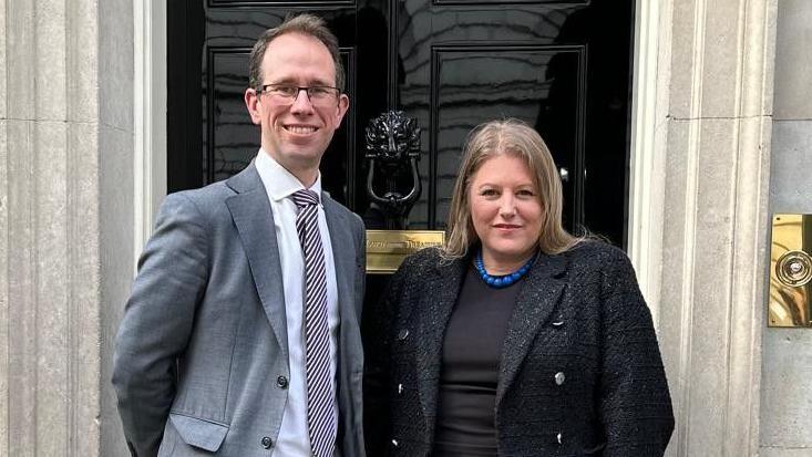 Matthew Barber and Donna Jones stand outside 10 Downing Street. Mr Barber is taller, has receding brown hair, and wears a light grey suit and glasses. Ms Jones has long, straight, fair hair, and wears a black jacket over a black top with a blue beaded necklace.