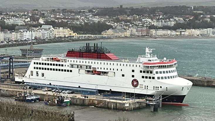 The Manxman ferry moored in Douglas Harbour. It is a large vessel painted in the Isle of Man Steam packet Company's colours of white, red and black, with the firm's logo featuring the three legs of Mann in a circle on the side. Douglas seafront can be seen in the background.