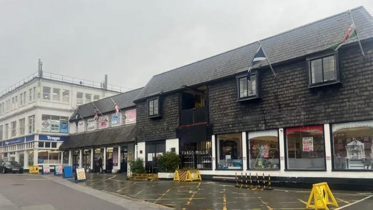 The two buildings which make up Trago Mills in Falmouth. One is wooden two-storey building and the other is a three-storey building along Arwenack Street. There is a hatched area in front of the wooden building and it has the nations' flags flying.