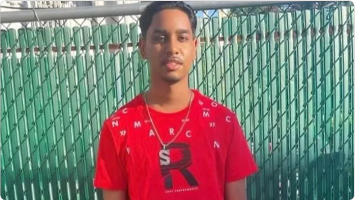 Shawn Seesahai wearing a red t-shirt and neck chain standing in front of green fence