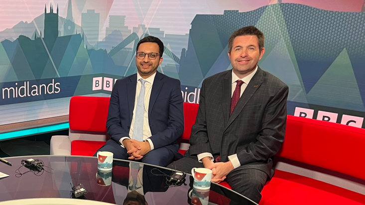Three people sit in a TV studio. A woman with long brown hair sits to the left in a red chair while two men in suits sit on a red sofa. A curved glass desk is positioned in front of them. 