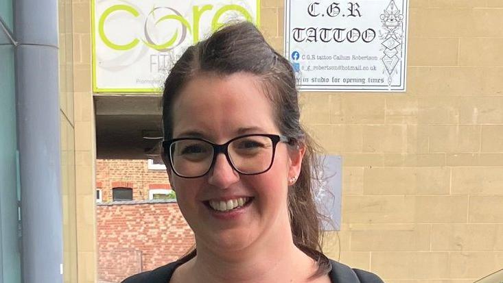 A young woman with tied back long brown hair and round black glasses smiles at the camera