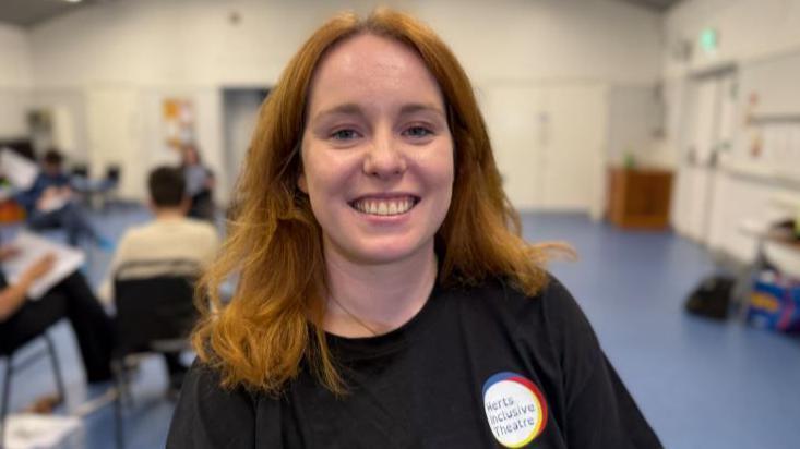 Kasia Truefitt has auburn shoulder-length hair and is smiling at the camera. She is wearing a black T-shirt with a Herts Inclusive Theatre badge.