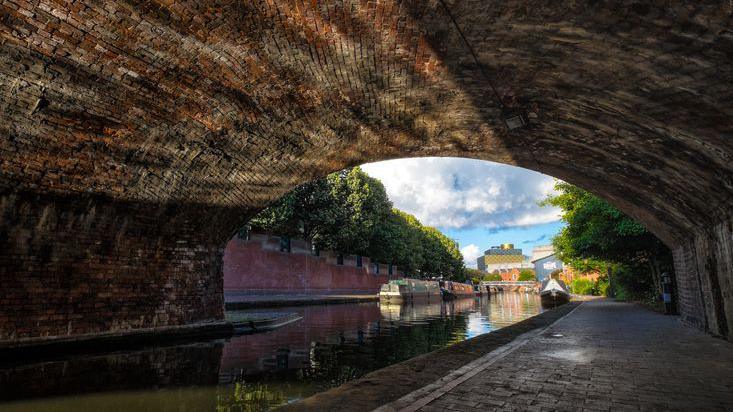 Light reflects on to the roof of a tunnel from a towpath on the Birmingham Canal Old Line. The city is visible in the distance and there is a view of the library building.