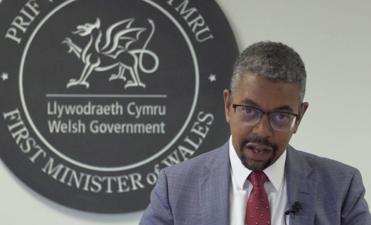 Vaughan Gething wearing a blue suit jacket with white shirt and red tie, he is wearing glasses and is in front of a Welsh government logo