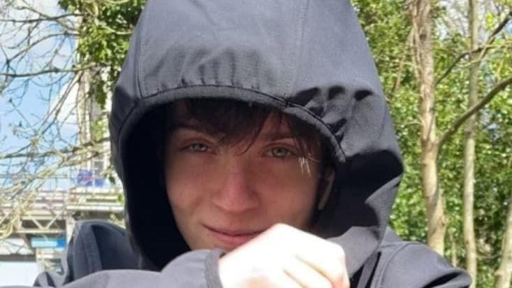 Daniel Davies smiling at the camera, sitting outdoors near some trees and wearing a black coat with his hood up. 