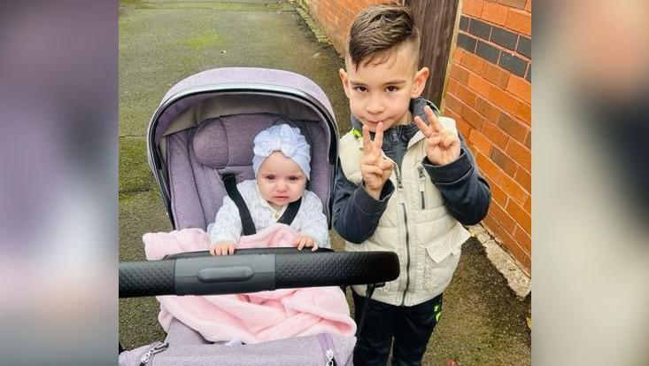 Sophia is in a pushchair and her brother is standing next to her. She is wearing a white hat with a bow and a white patterned top and is covered by a pink blanket. Her brother is wearing a cream gilet and dark top and trousers. They are on a pavement with a brick wall seen to the right of them.