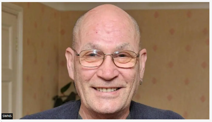 A bald man wearing a dark top and glasses. He has an earring in his left ear.