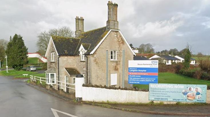 Google Street View image of the entrance to the Langdon Hospital site in Dawlish, Devon. A house which is part of the site is next to the junction of the road where the entrance is. A blue, white and red sign with the hospital's name on it along with other directional information is next to a white stone wall. A sign with a recruitment advert on it is also on the wall.