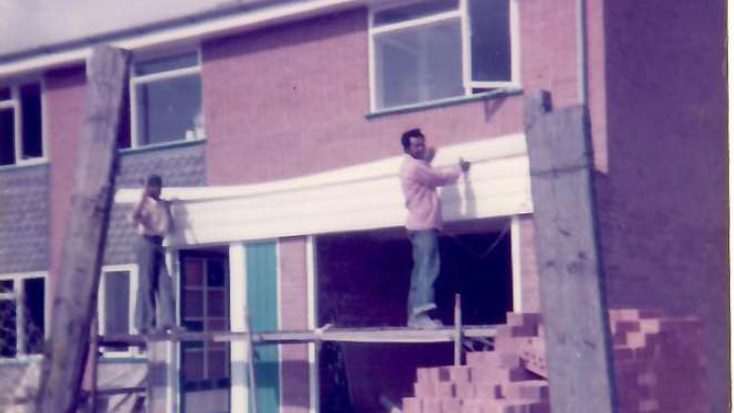 An old image of two men fixing up the front of a house. There are pieces of wood and piles of bricks in the foreground.